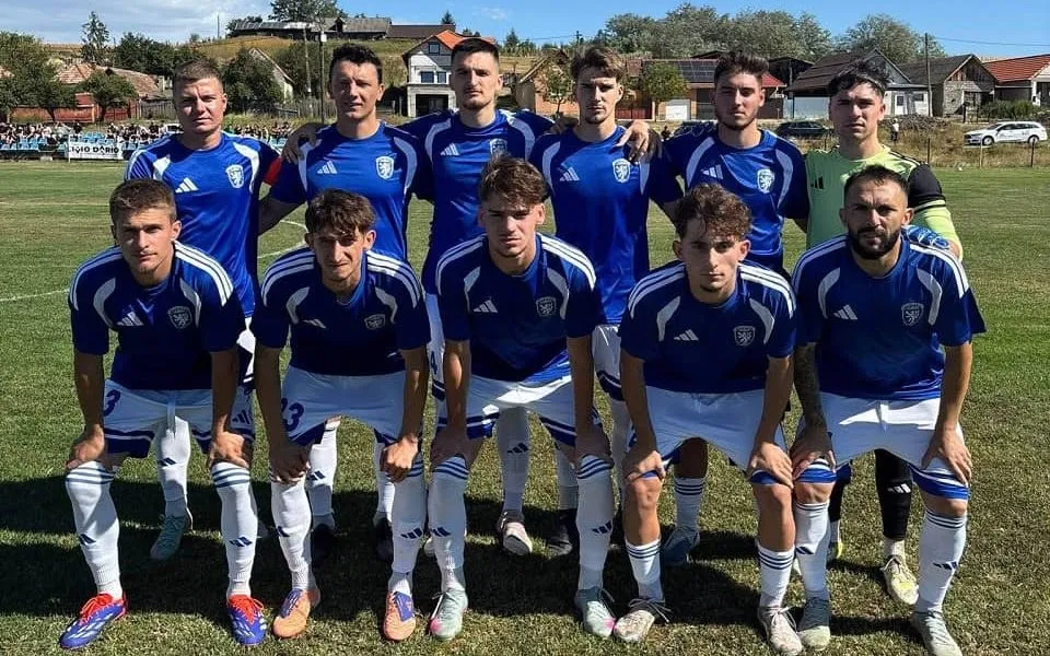 Unsprezece jucători în tricouri albastre și pantaloni scurți se aliniază pe un teren de fotbal, șase în picioare și cinci aplecați în față.