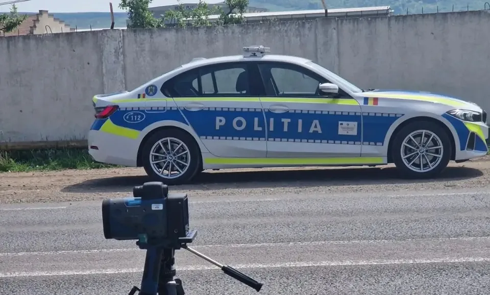 În prim-plan se află un aparat negru pe trepied, iar în spate este o mașină albă de poliție, lângă un zid.
