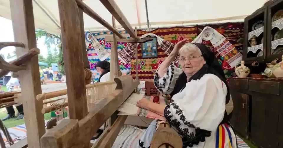 O femeie în vârstă stă lângă un război de țesut din lemn, cu mâinile pe fire, într-o încăpere cu țesături colorate.