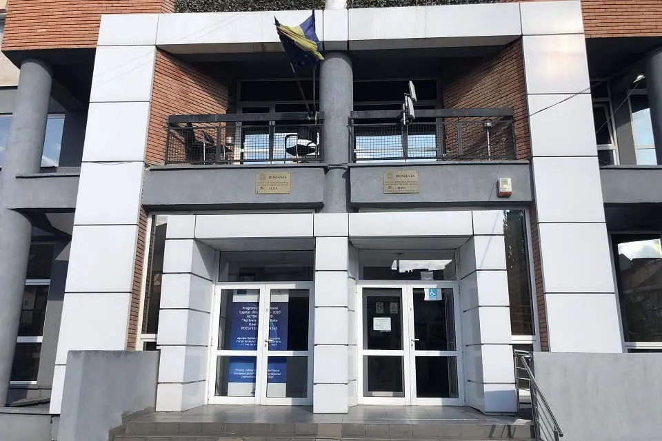 Fațada unei clădiri are coloane gri, balustradă metalică, două intrări vitrate, afișe albastre pe geamuri și un steag deasupra balconului.