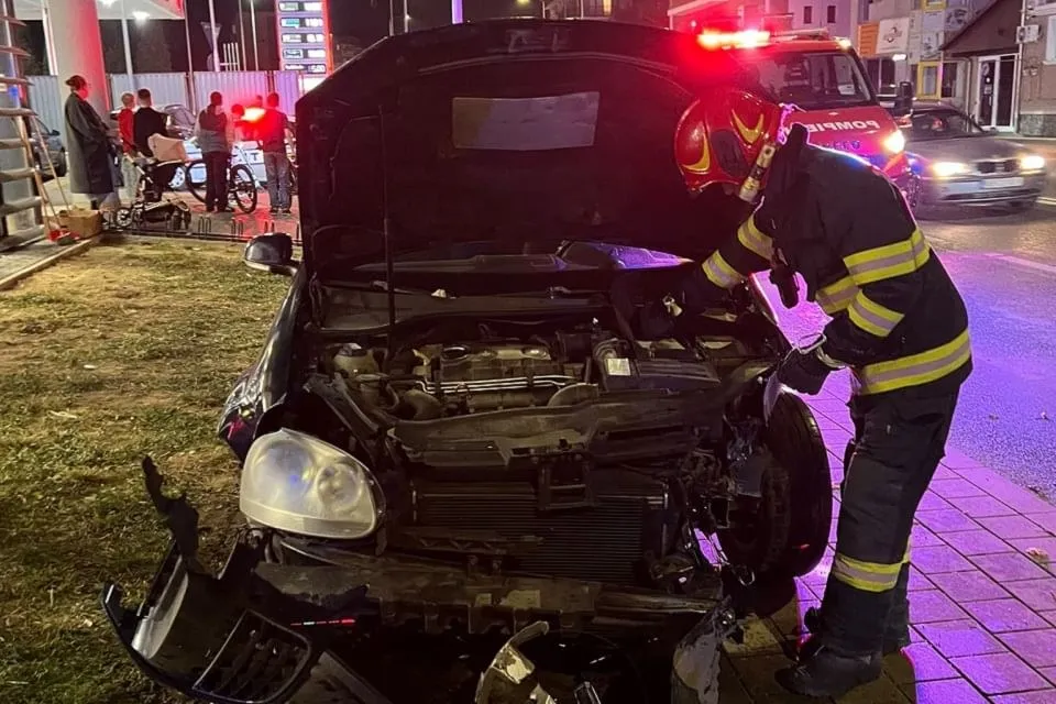 Un autoturism cu partea frontală avariată și capota ridicată se află lângă o persoană în uniformă reflectorizantă; în spate apar vehicule și oameni.