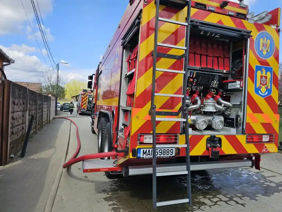 Două autospeciale roșii sunt oprite pe o stradă îngustă, iar din vehiculul apropiat coboară un furtun și o scară metalică.