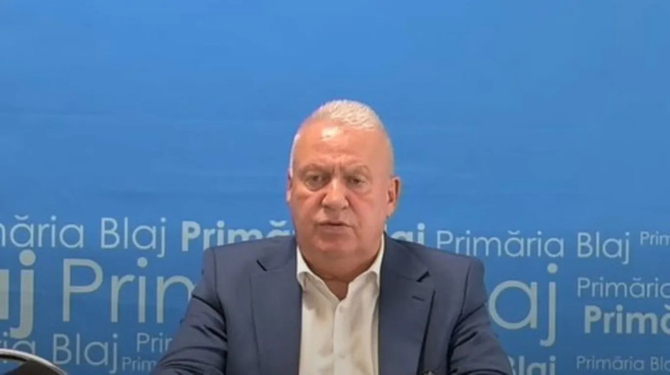 Un bărbat cu păr alb, îmbrăcat într-un sacou bleumarin și o cămașă albă, stă în fața unui fundal albastru cu inscripții.