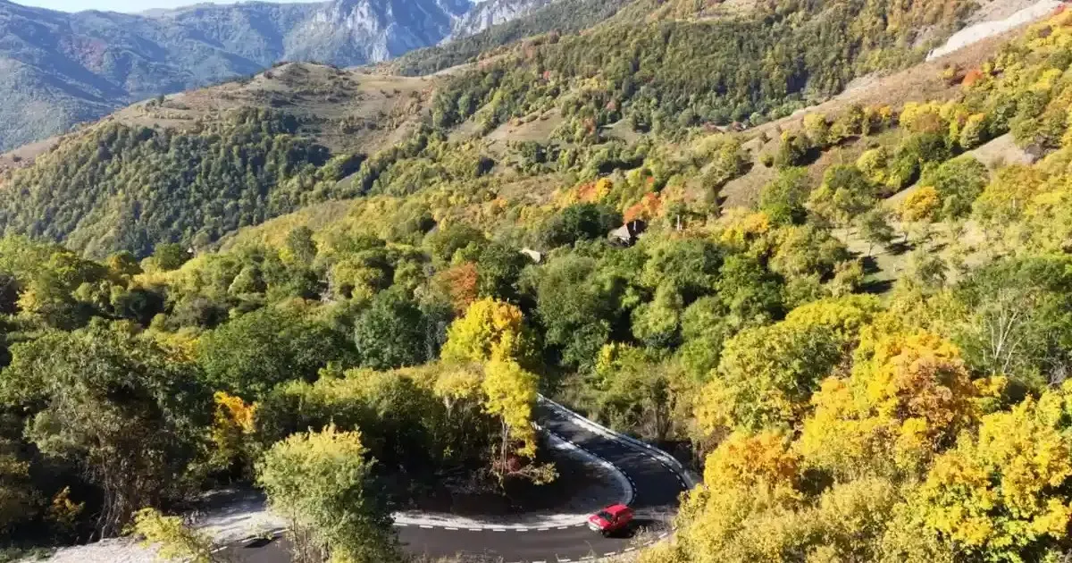 Un drum asfaltat șerpuitor traversează o vale împădurită, iar un vehicul roșu se află pe curbă.