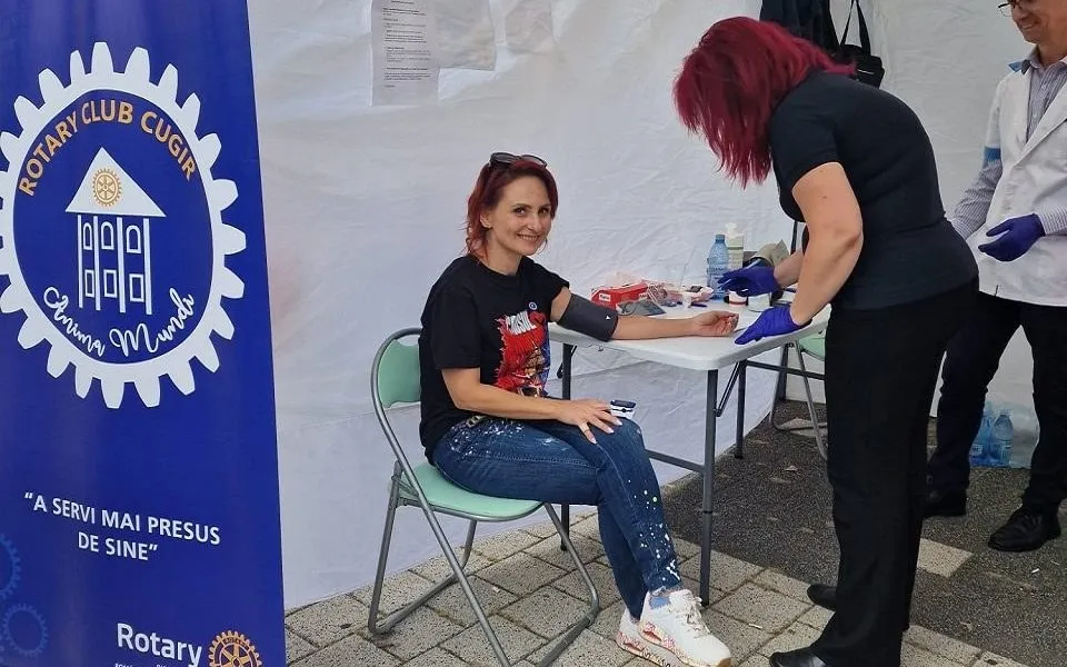 O femeie așezată ține brațul pe masă, iar două persoane în picioare poartă mănuși; alături se află un banner albastru.