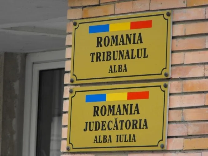 Două plăci galbene cu text negru și benzi tricolore sunt fixate pe un perete de cărămidă, lângă o ușă.