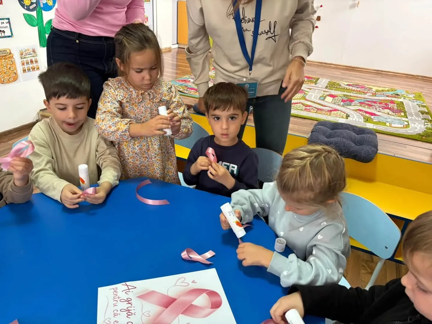 Patru copii stau în jurul unei mese albastre, ținând hârtie și obiecte mici; pe masă se văd fundițe roz.