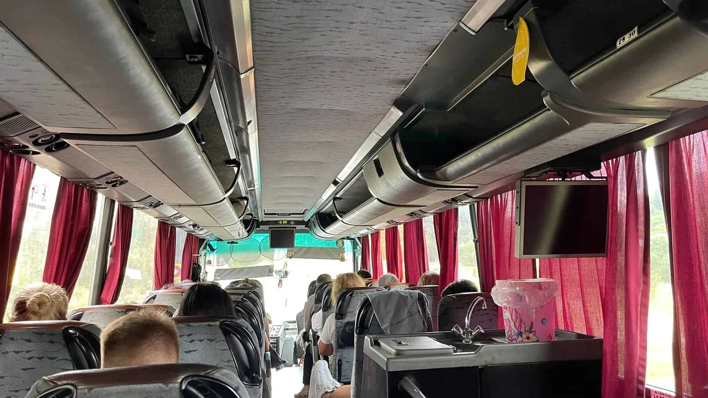 Interiorul unui autocar prezintă pasageri așezați, scaune gri, compartimente suspendate, perdele roșii, un ecran și o cutie transparentă în prim-plan.