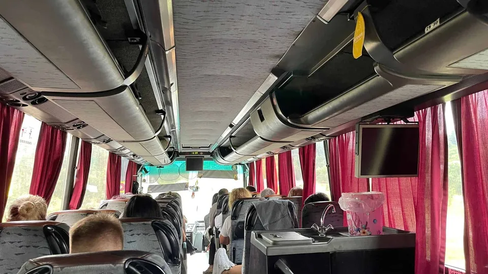 Interiorul unui autocar prezintă pasageri așezați, scaune gri, compartimente suspendate, perdele roșii, un ecran și o cutie transparentă în prim-plan.