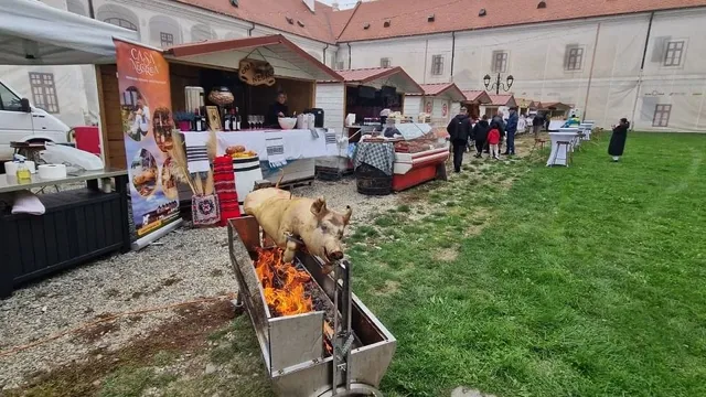 Un porc este așezat pe o frigare deasupra unui foc, lângă corturi cu mese și câteva persoane, într-o curte cu iarbă.