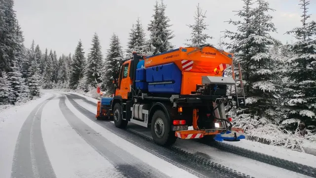 Un camion portocaliu și albastru circulă pe un drum acoperit parțial de zăpadă, printre copaci înzăpeziți; în spatele lui se văd dâre întunecate.