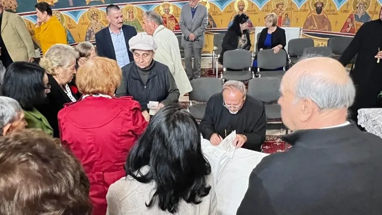 Mai multe persoane sunt adunate în jurul unei mese, iar un bărbat așezat scrie într-o carte deschisă.