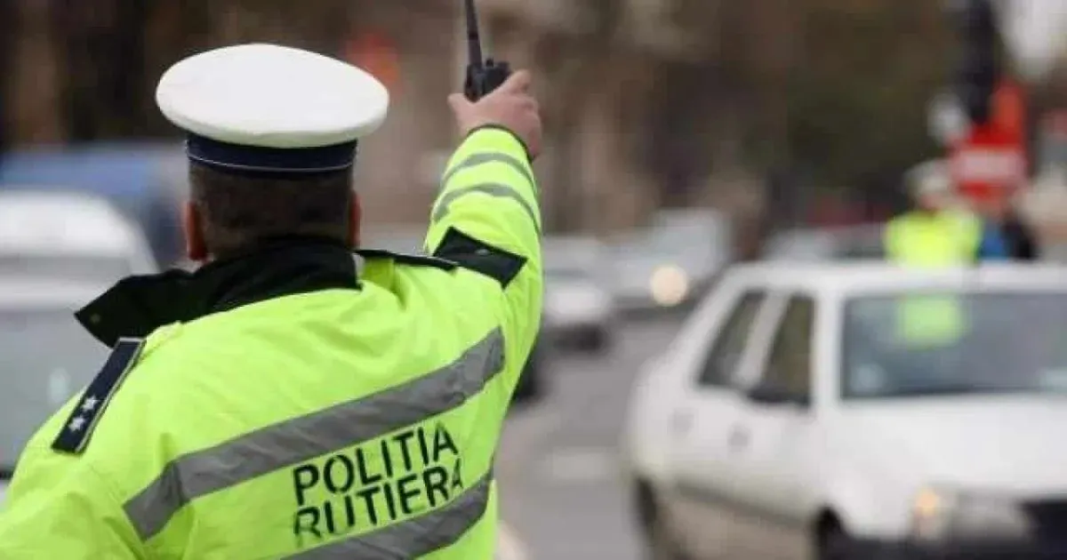 Un bărbat în uniformă fluorescentă ridică brațul lângă un autoturism alb, pe o stradă.