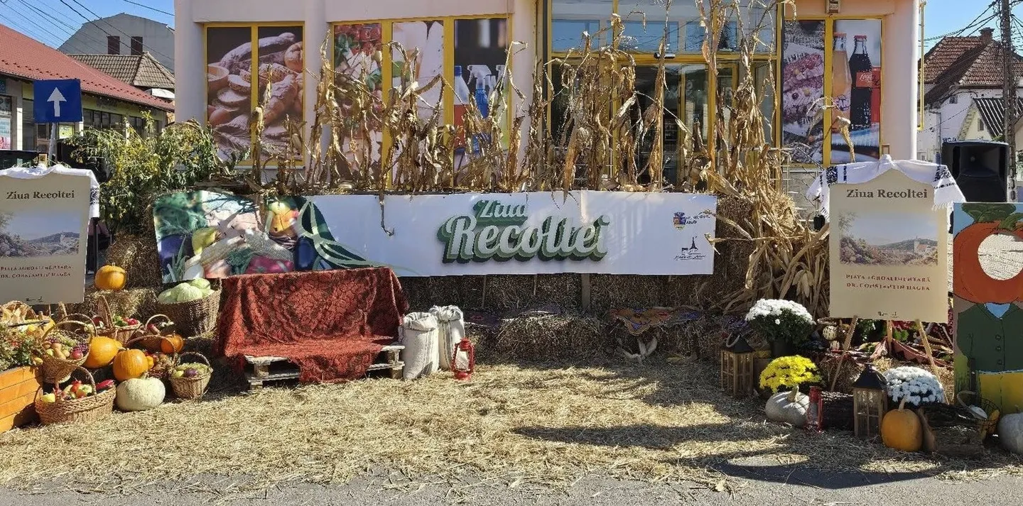 Un decor cu coceni de porumb, baloți de paie, dovleci, coșuri cu legume, crizanteme, ulcioare și o bancă acoperită cu țesătură.