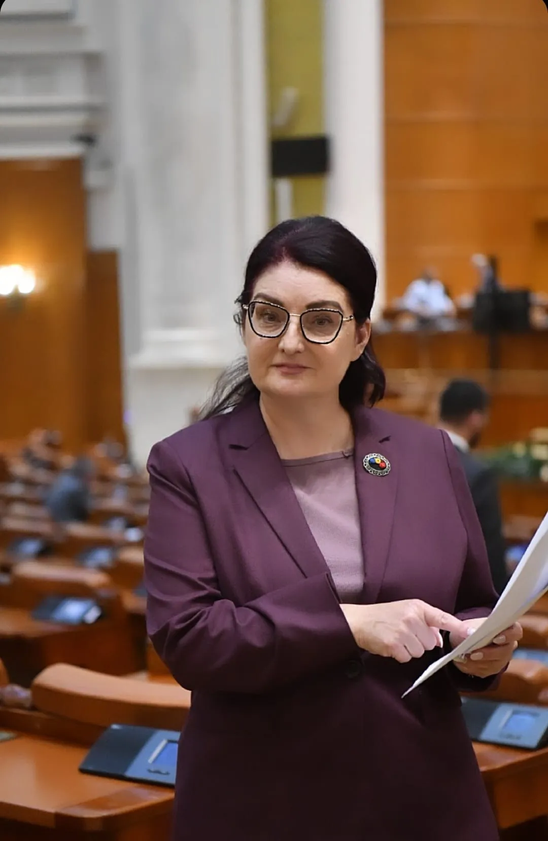O femeie cu ochelari și sacou mov ține câteva foi în fața unor rânduri de bănci din lemn.