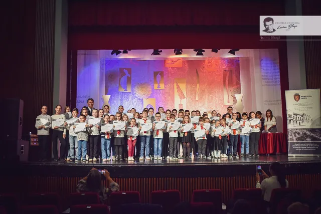 Un grup numeros de copii și adulți stă pe o scenă, majoritatea ținând foi albe, în fața unui decor iluminat colorat.