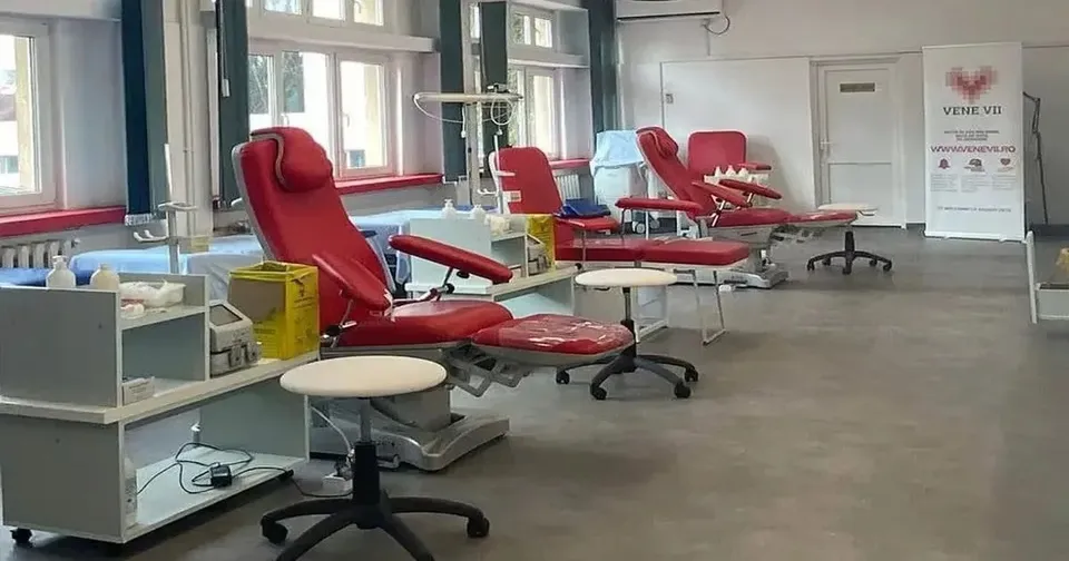Trei fotolii roșii înclinate sunt aliniate lângă ferestre, între cărucioare cu recipiente, suporturi metalice și scaune rotunde, într-o încăpere cu podea gri.