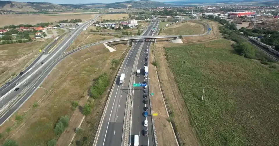 Vedere aeriană a unei autostrăzi și a unor bretele rutiere, cu vehicule, un pasaj, terenuri cu iarbă și clădiri în depărtare.