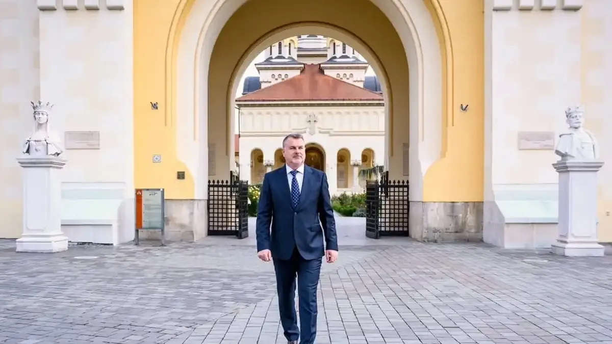Un bărbat în costum bleumarin și cravată merge pe pavaj, în fața unei arcade; în spate se văd o clădire și două statui.