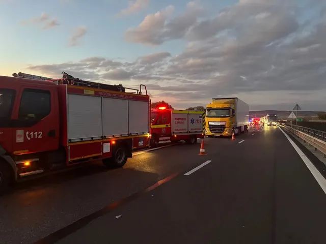 Pe o șosea se văd un camion roșu cu compartimente laterale, o ambulanță și un camion galben, lângă conuri portocalii și balustrade metalice.