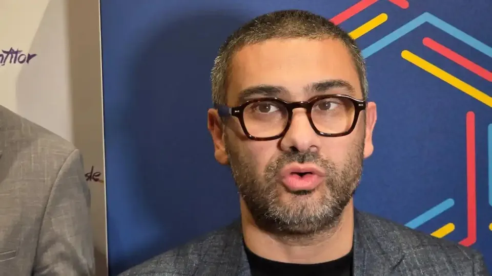 Un bărbat cu ochelari negri, barbă scurtă și sacou gri apare în prim-plan, în fața unui panou albastru cu linii colorate.