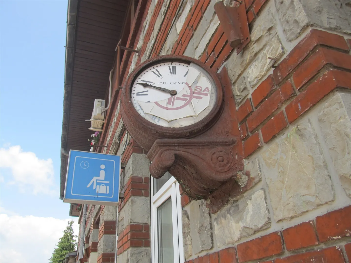 Un ceas rotund cu cifre romane este montat pe un perete de cărămidă și piatră, lângă un indicator albastru.