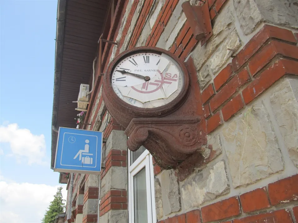 Un ceas rotund cu cifre romane este montat pe un perete de cărămidă și piatră, lângă un indicator albastru.