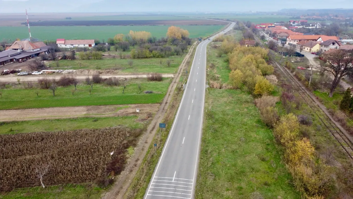 Un drum asfaltat traversează câmpuri agricole, având o cale ferată și case cu acoperișuri roșii în partea dreaptă.