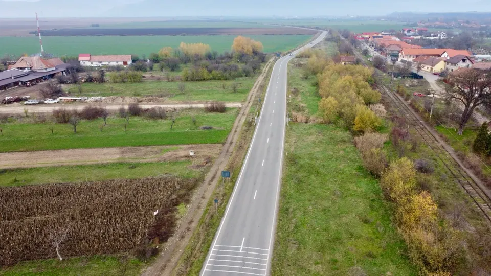 Un drum asfaltat traversează câmpuri agricole, având o cale ferată și case cu acoperișuri roșii în partea dreaptă.