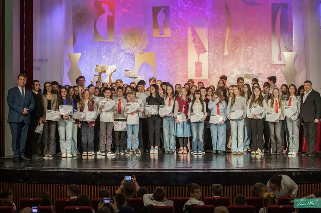 Un grup numeros de tineri și adulți stă pe o scenă, mulți ținând diplome, iar spectatori fotografiază din fotolii.