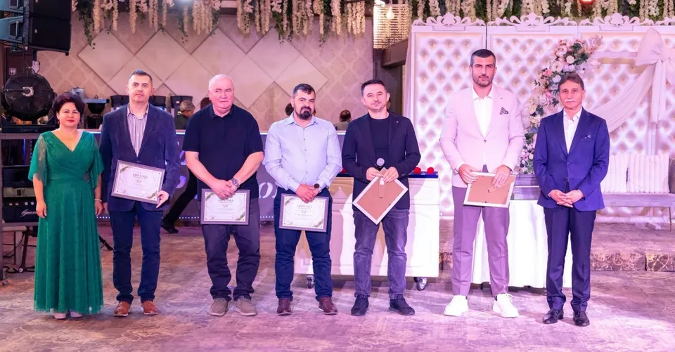 Șase bărbați și o femeie stau aliniați într-o sală decorată; cinci țin mape sau diplome, iar în spatele lor se află o masă albă.