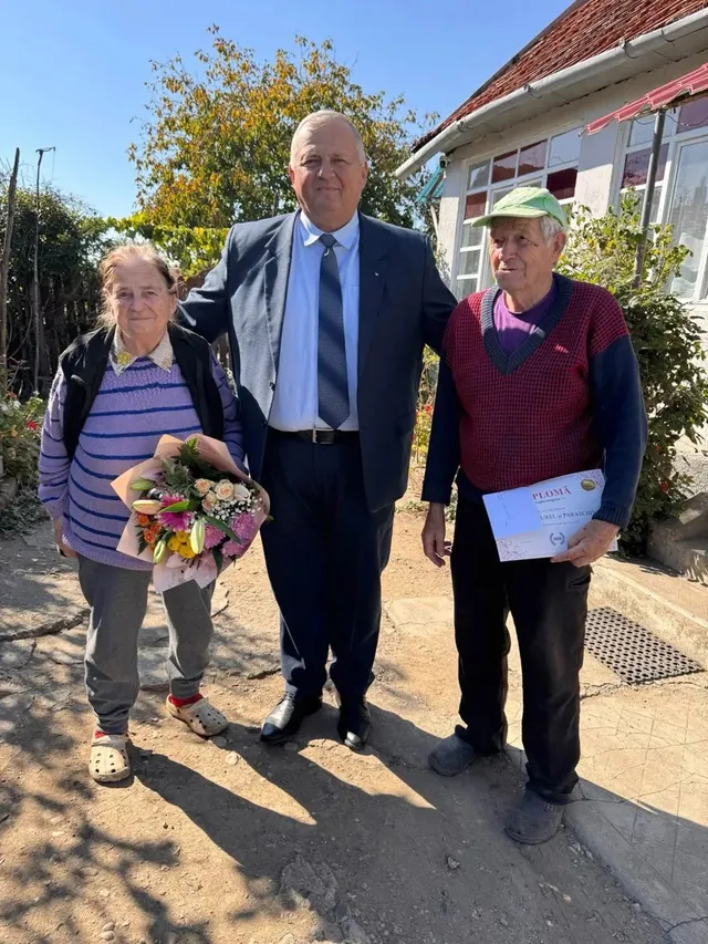 Trei persoane stau în fața unei clădiri; femeia din stânga ține un buchet, iar cea din dreapta ține o foaie cu text.