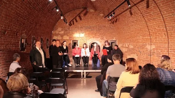 Mai multe persoane stau pe scaune sau în picioare într-o încăpere cu pereți și tavan boltit din cărămidă, în fața unor obiecte expuse.