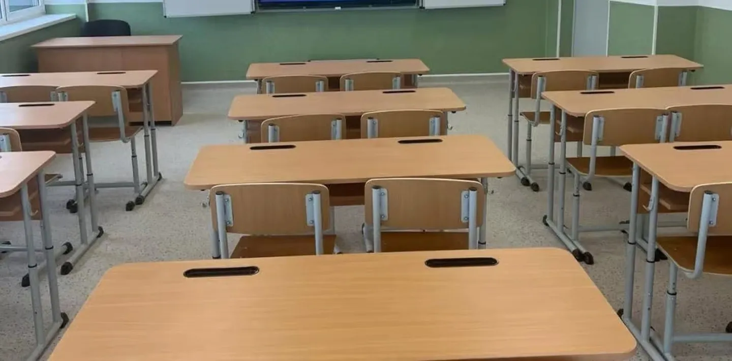 Într-o sală sunt aliniate mese individuale cu blaturi din lemn și scaune, iar în față se vede o tablă verde.