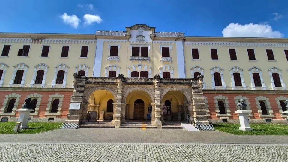 O clădire monumentală galbenă și albă, cu trei arcade la intrare, numeroase ferestre și o piață pavată, apare sub cerul albastru.