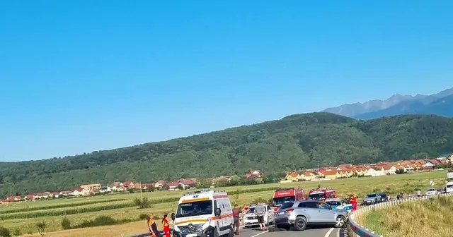 Pe un drum de lângă câmpuri se văd o ambulanță, mai multe mașini oprite și câteva persoane, cu dealuri în fundal.