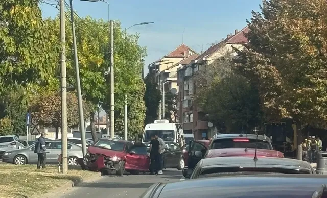 O mașină roșie avariată stă lângă mai multe vehicule, cu câțiva oameni în jur, pe o stradă mărginită de copaci și clădiri.