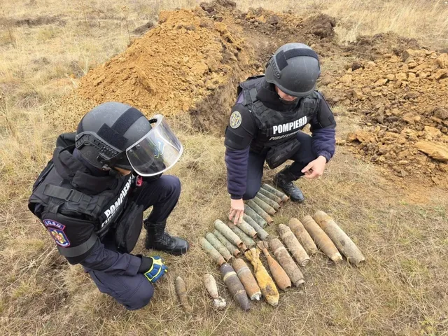Două persoane în uniforme închise la culoare și căști stau lângă mai multe obiecte metalice alungite, așezate pe pământ.