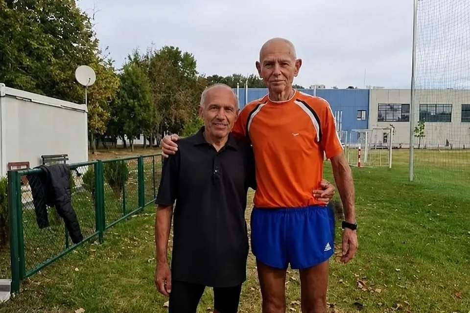 Doi bărbați stau alături pe iarbă, lângă un gard; unul poartă haine negre, iar celălalt un tricou portocaliu și pantaloni scurți albaștri.