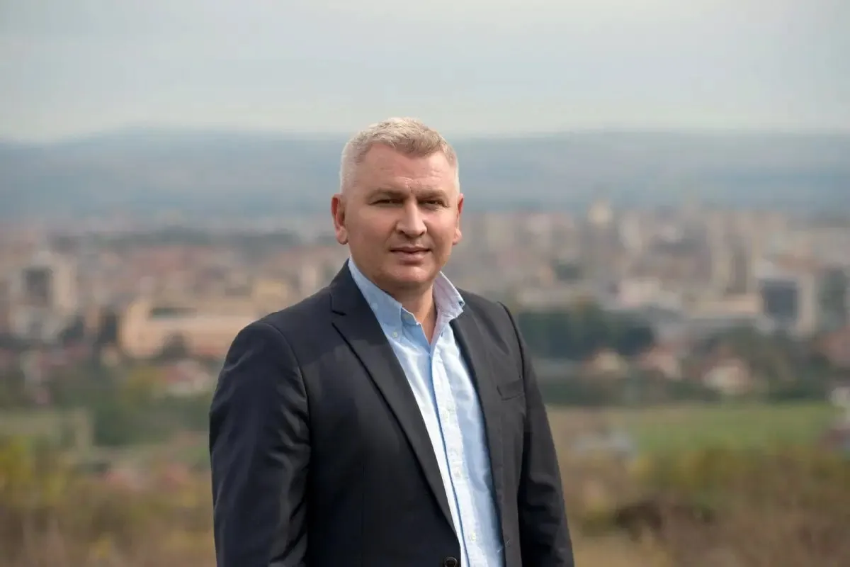 Un bărbat cu păr scurt, cărunt, poartă un sacou închis la culoare și o cămașă deschisă la culoare, în fața unui fundal urban neclar.