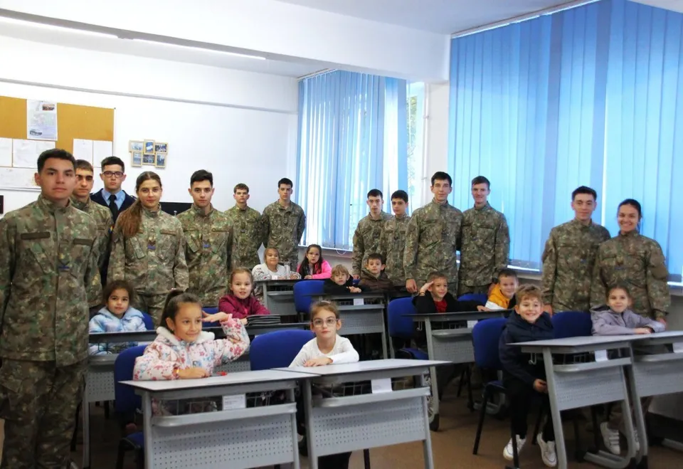 Copii stau la bănci într-o sală de clasă, iar tineri în uniforme camuflate sunt aliniați în spatele lor și lângă bănci.