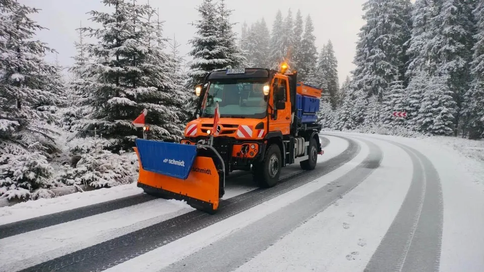Un utilaj portocaliu cu lamă albastră este pe un drum acoperit parțial de zăpadă, mărginit de copaci înzăpeziți.