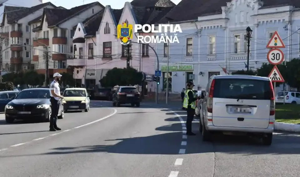 Doi polițiști rutieri stau pe o stradă lângă mai multe autoturisme, clădiri și indicatoare, iar un autovehicul alb este oprit pe dreapta.