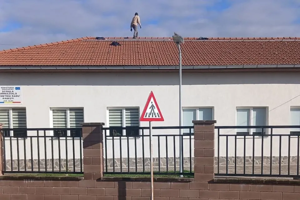 O persoană se află pe acoperișul roșcat al unei clădiri albe; în față sunt un gard metalic și un indicator rutier triunghiular.