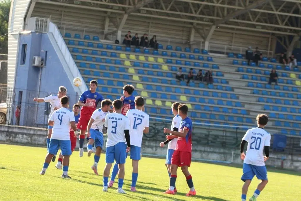 Mai mulți jucători în tricouri albe și albastre și alții în tricouri roșii sunt grupați pe un teren de fotbal, sub tribune cu scaune albastre.