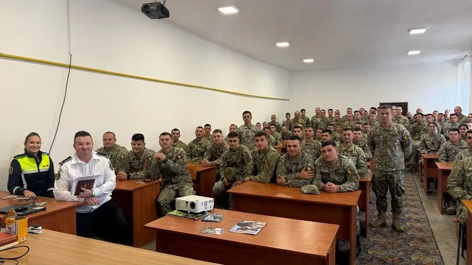Într-o sală, două persoane stau în fața unor rânduri de persoane în uniforme militare, așezate la bănci; pe bănci sunt hârtii și un proiector.
