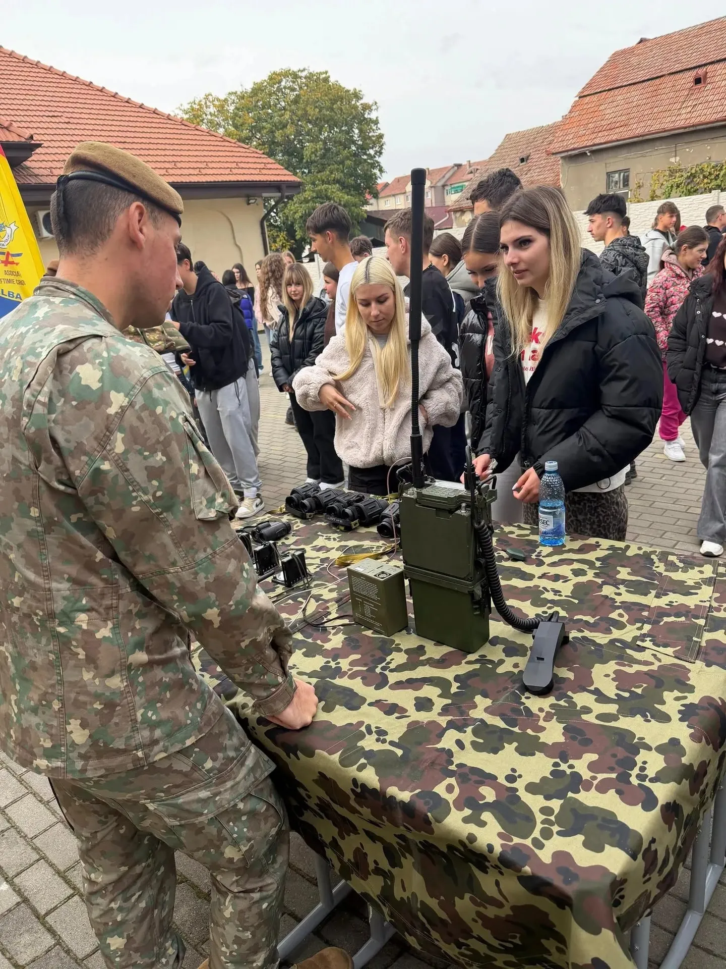 Un bărbat în uniformă camuflată stă lângă o masă acoperită cu material camuflat, pe care se află echipamente, în fața unui grup de tineri.
