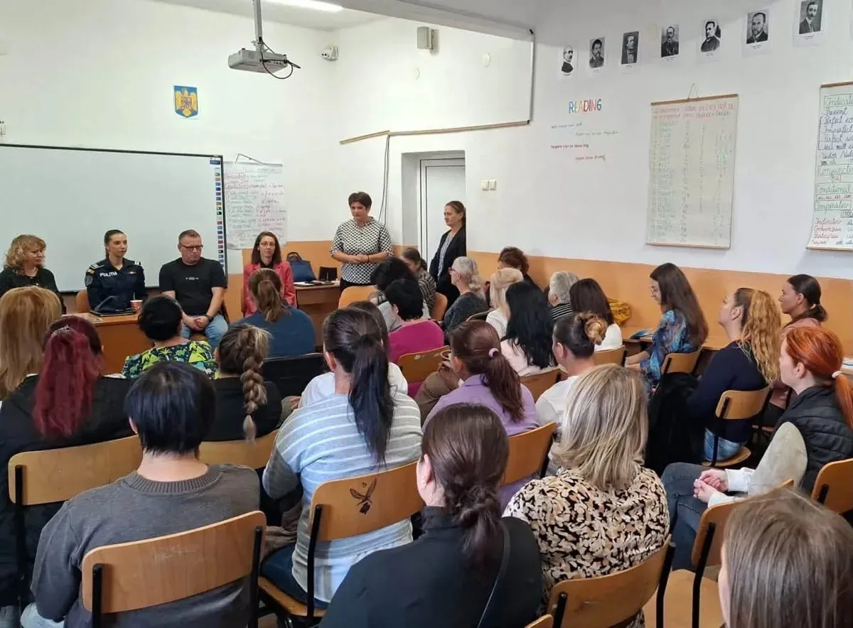 Mai multe persoane stau pe scaune într-o sală de clasă, în timp ce câteva persoane sunt așezate sau stau în picioare în față.