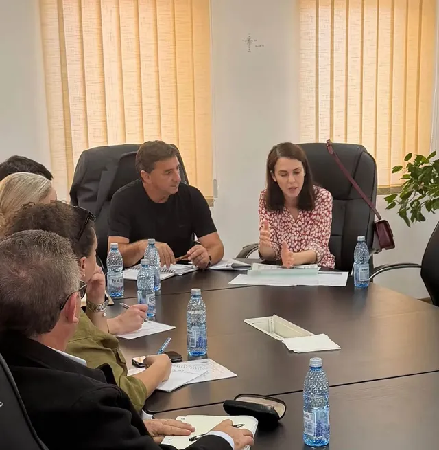 Mai multe persoane stau în jurul unei mese de conferință; două sunt așezate în capătul mesei, cu documente și sticle de apă în față.