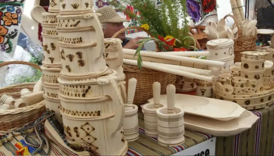 Pe o masă acoperită cu țesătură sunt expuse vase ceramice decorate, farfurii, suporturi cilindrice, coșuri împletite și obiecte textile colorate.